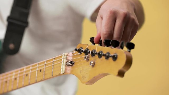 Cropped photo of young male guitarist in casual clothing fixing tuning pegs on electric guitar closeup, isolated over yellow background