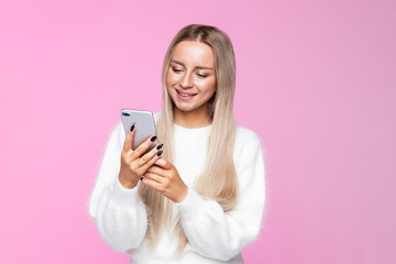 Cheerful young European blonde female smiling and using mobile phone, talking on video call, dressed in fluffy white sweater, pink background/
