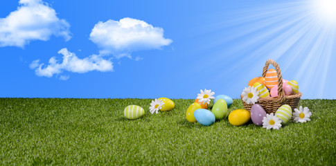 basket of multi colored painted easter eggs on green grass with springtime daisy flowers and blue sky