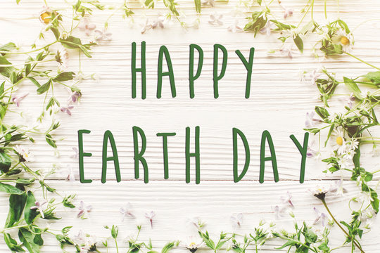 happy earth day text sign on beautiful lilac and daisy flowers on rustic white wooden background top view. greeting card. environmental concept. earth day. save environment. eco