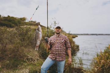 Young handsome unshaven man in checkered shirt, cap and sunglasses holds a fishing pole with caught...