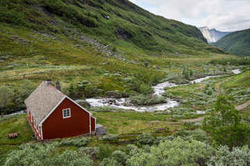 Fototapeta na wymiar Red wooden cottage next to a mountain stream in a valley during a cloudy day near the Myrdal railway station in Norway