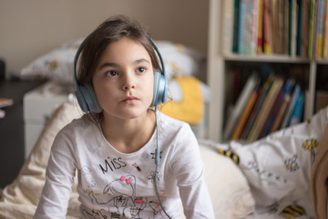 the girl is sitting in bed with headphones. Behind her is a shelving with children's books