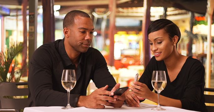 An African American couple use their mobile phones while eating dinner. A black man and woman use their smart devices while drinking wine