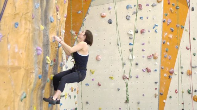 On climbing woman is hooked on wall in gym during exercise. With an effort female sports person overcomes artificial obstacles during sports practice for courage and endurance.