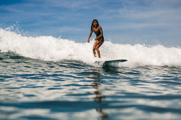 Beautiful young indonesian woman in bikini surfing wave in Bali on the background of blue sky, clouds and tropical beach