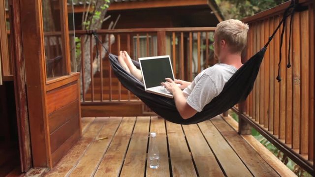 man working behind a laptop and drinks water in a hammock on a terrace