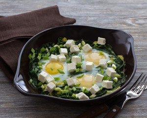 Green shakshuka with spinach, leek and feta in a ceramic frying pan on a wooden table with bread, selective focus. Delicious healthy home food