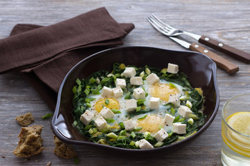 Green shakshuka with spinach, leek and feta in a ceramic frying pan on a wooden table with bread, selective focus. Delicious healthy home food