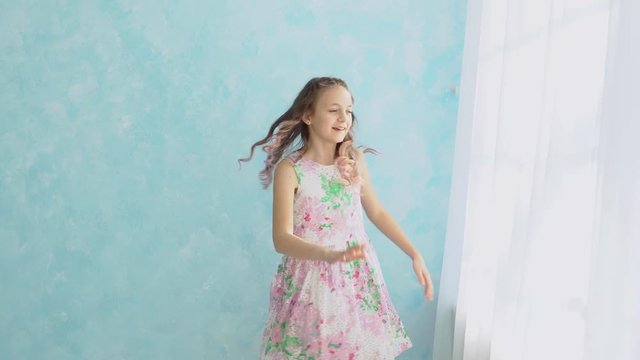 Girl in a beautiful floral dress is dancing against the background of a blue wall next to the window.