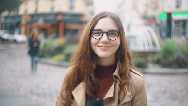 Portrait of young beautiful french student in glasses standing on the street, drink tea from cap and smiling at camera.