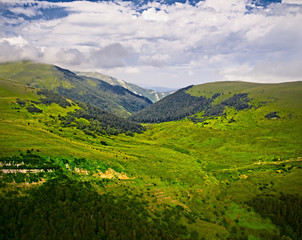Obraz premium Aerial view of green hills. Summer landscape. Green grassy meadow on a hillside on top of mountain ridge with some forest under cloudy blue sky. Adygea, Lago-naki, Lagonaki, Krasnodar, Russia.
