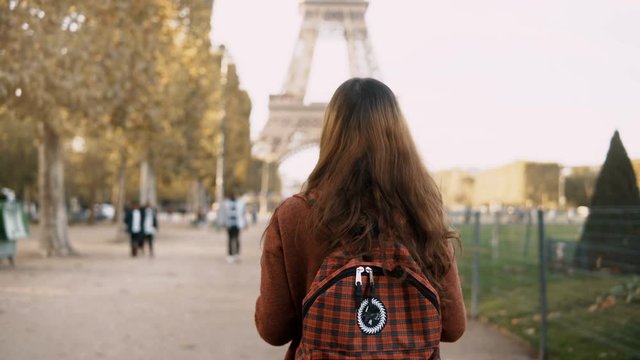 Back view of young woman walking with backpack near the Eiffel tower in Paris, France. Tourist enjoying the view.