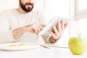 Portrait of blurry adult man in casual shirt having healthy meal with frying eggs in apartment, while using digital tablet