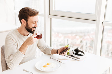 Happy mature guy drinking tea and using mobile phone, while having healthy lunch with frying eggs in hotel room