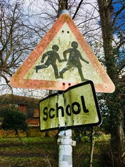Weathered grunge and moss covered school kids crossing signs on a pole with winter bare trees in the background against a cloudy blue sky. Reigate, England