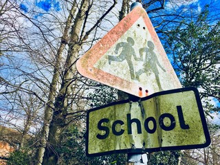 Weathered grunge and moss covered school kids crossing signs on a pole with winter bare trees in the background against a cloudy blue sky. Reigate, England