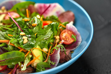 Spicy beef slices meat salad with tomatoes, parsley, radish and salad leaves spinach in a blue plate on a black table. Close-up