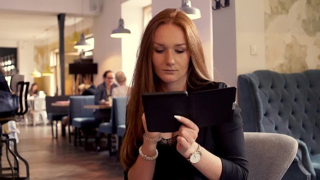 Beautiful woman with red hair reading something on e-book sitting in cafe 
