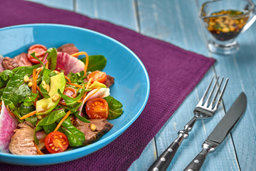 Spicy beef slices meat salad with tomatoes, parsley, radish and salad leaves spinach in a blue plate on a wooden table.