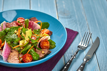 Spicy beef slices meat salad with tomatoes, parsley, radish and salad leaves spinach in a blue plate on a wooden table.