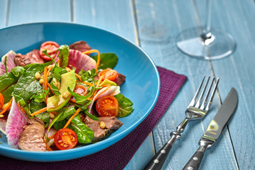 Spicy beef slices meat salad with tomatoes, parsley, radish and salad leaves spinach in a blue plate on a wooden table.