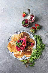 Homemade pancakes served on decorate plate with berries, mint and fruit salad inside pink dragon fruit, glass jar of yogurt over grey texture background. Top view, space.