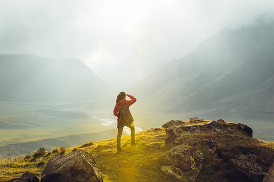 Discovery Travel Destination Concept. Hiker Young Woman With Backpack Rises To The Mountain Top Against Backdrop Of Sunset, Rear View