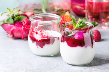 Jars of natural white yogurt with berry sauce, fruit salad with pink dragon fruit, berries and mint, served with bottle of lemonade on grey table. Close up. Healthy eating