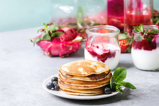 Stack of homemade pancakes served on plate with berries, mint, glass jars of yogurt, bottle of lemonade, fruit salad in pink dragon fruit over grey texture table.
