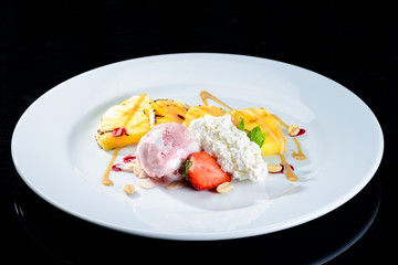 Sweet appetizing dessert with ice-cream, pineapple, strawberries, nuts and mint in a white plate on dark background