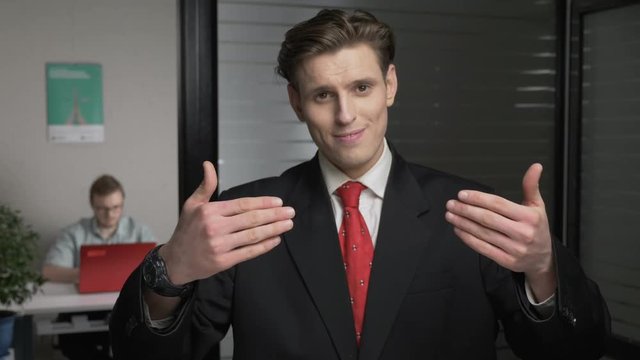 Young successful businessman in suit showing a come here sign. Man works on a computer in the background. 60 fps