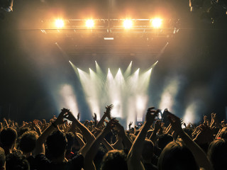 Concert crowd attending a concert, people silhouettes are visible, backlit by stage lights. Raised hands and smart phones are visible here and there.