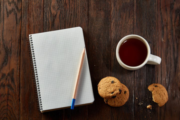 Cup of tea with cookies, workbook and a pencil on a wooden background, top view