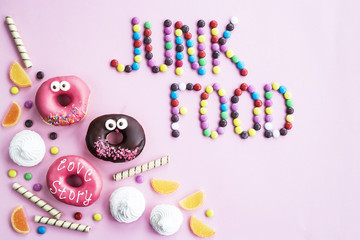 The phrase Junk food is lined with chocolate glazed balls on a pink background. Next to donuts, marmalade, chocolate sticks and meringue. Unhealthy food concept. Top view