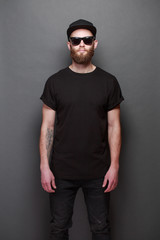 Obraz premium Hipster handsome male model with beard wearing black blank t-shirt with space for your logo or design over gray background