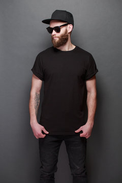 Hipster handsome male model with beard wearing black blank t-shirt with space for your logo or design over gray background