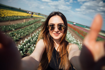 Fototapeta premium Young beautiful woman wear sunglasses make selfie in tulips flower field in sunny spring day