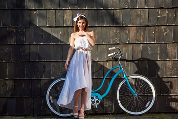 Lovely girl on a sunny day against a blue vintage bicycle and a dark wall. The girl is dressed in light clothes and looking at the camera with a smile