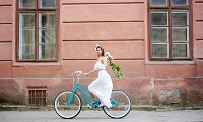 Fototapeta premium Smiling woman on a vintage bicycle is riding along the old city with peonies in her hand. The girl is dressed in a light dress and an accessory on her head