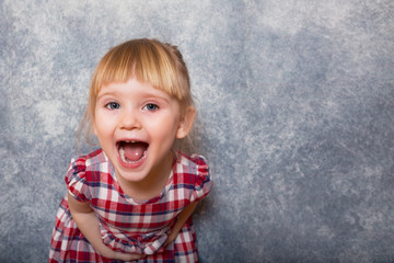 A tiny sweet girl with blond hair wearing a red checkered dress. She makes faces on a gray...