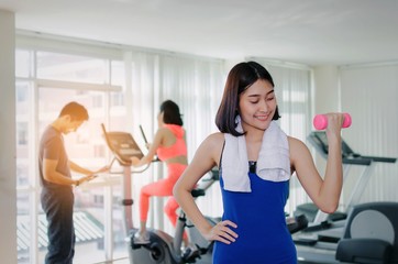 young asian pretty woman doing exercises with dumbbell at biceps in fitness gym with people background, bodybuilder, healthy lifestyle, exercise fitness, workout and sport training concept, soft focus