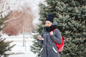 Modern girl hipster listening to music on headphones, smiling and looking at mobile phone outdoors. Portrait of a beautiful girl with a phone in hands.