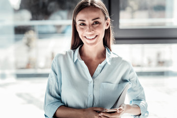 Modern igen. Joyful happy nice woman smiling and looking at you while holding her smartphone