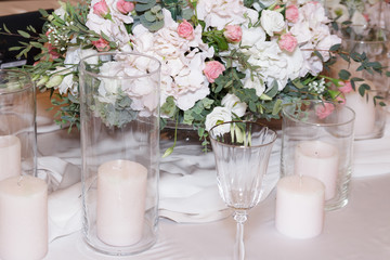 On the table, decorated for the wedding ceremony in the stylish restaurant, a colored candle, white dishes, glasses, bouquets with fresh flowers and modern wedding decor stand on tablecloth. Side view