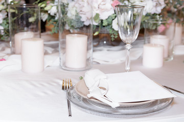 On the table for the stylish wedding ceremony in the restaurant, a colored candle, white dishes, glasses, bouquets with fresh flowers and wedding decor stand on a light tablecloth. Selective focus