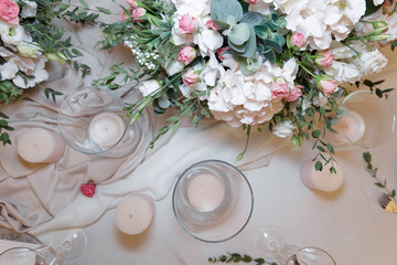 On the table, decorated for the wedding ceremony in the restaurant, a colored candle, white dishes, glasses, bouquets with fresh flowers and modern wedding decor stand on a silk tablecloth. Top view