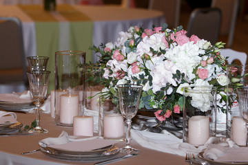 A stylish wedding decor of dishes, white bouquets with flowers, many candles and glasses stand on a light tablecloth of a served table in a restaurant. Side view, selective focus