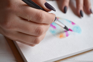 Woman paints in a sketchbook. Tools for painting on the white wooden desk. Painter work.