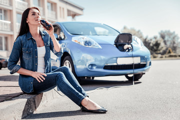 So hot. Young cute pleasant woman charging the electric car drinking cola on the board and looking aside.